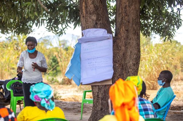 A Kenyan woman leads an outdoor session under a tree where a flip chart hangs while seated people listen.