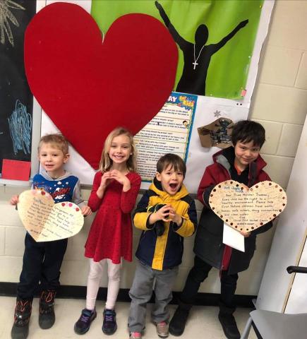 Three children around 5-7 years old stand in a church hallway holding up heart-shaped letters they wrote to the prime minister.