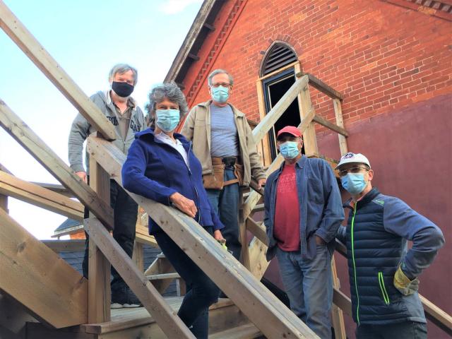 A group of four men and one women stand on the temporary stairs outside their church building to observe "greening" construction progress. They are wearing pandemic masks.
