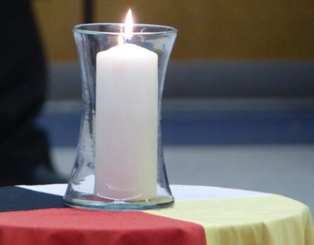 Burning Tall white candle in clear glass jar sits on a cloth with the four Indigenous colours of black, white, yellow, and red.