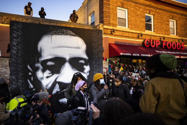 A crowd stands before a George Floyd mural at the spot where he was murdered by police office Derek Chauvin.