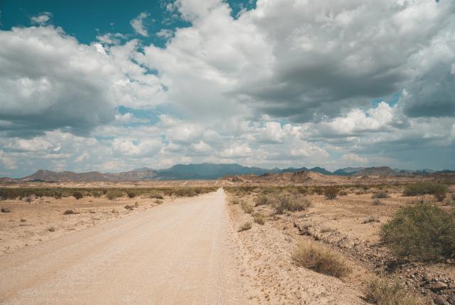 A dirt road running straight through the sparse desert, with dramatic clouds overhead.