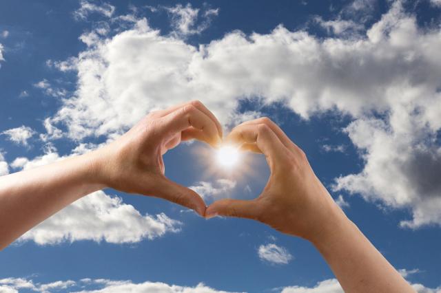 Two hands form a heart against a backdrop of a blue sky with white clouds.
