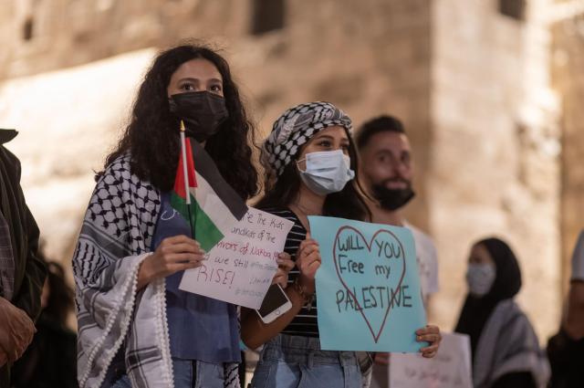 Two Palestinian women hold up candles and signs at a protest. One of the signs reads Will You Free My Palestine.