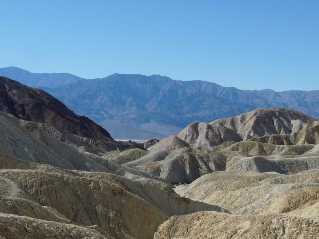 A desert scene with rugged yellow rock outcrops in the foreground, and a purple mountain range in the distance. Lots of blue sky.