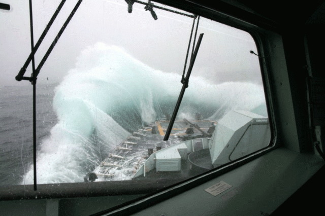 A giant wave hits the front of a Navy ship, slashing over it.