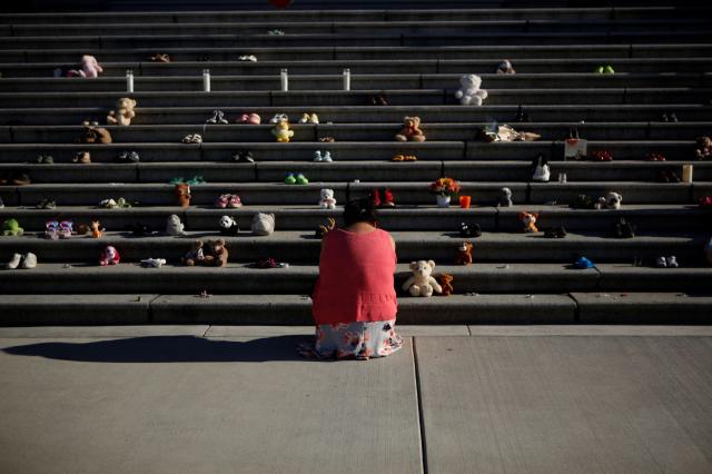 A woman dressed in orange kneels in front of the Victoria legislature steps, where children's shoes have been lined up.