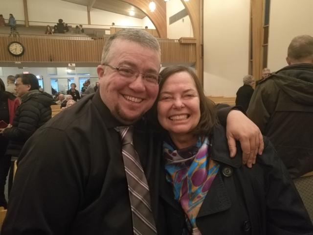 The author, Jordan Sullivan, a man with short light coloured hair, glasses, and a light beard, wraps his arm about his friend, the choir director, who is a woman of about the same age, with dark hair. Both are dressed for a fancy occasion and have terrific smiles!