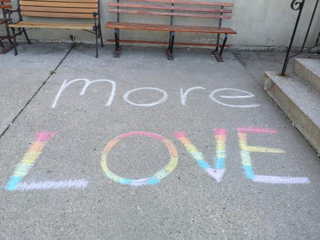 The words "More Love" written on a sidewalk in rainbow chalk colours.