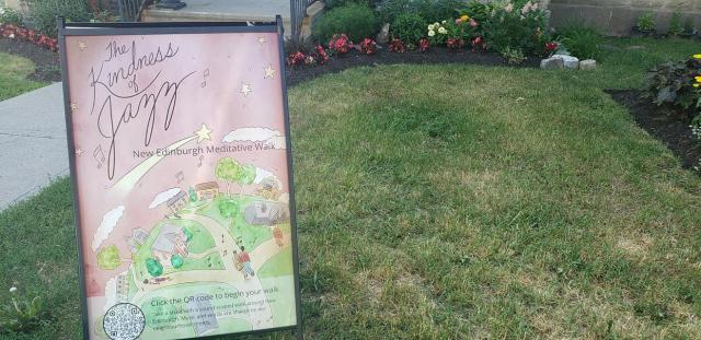 A signboard holding a poster with the words The Kindness of Jazz on a background illustration of people walking paths is displayed on a lawn.