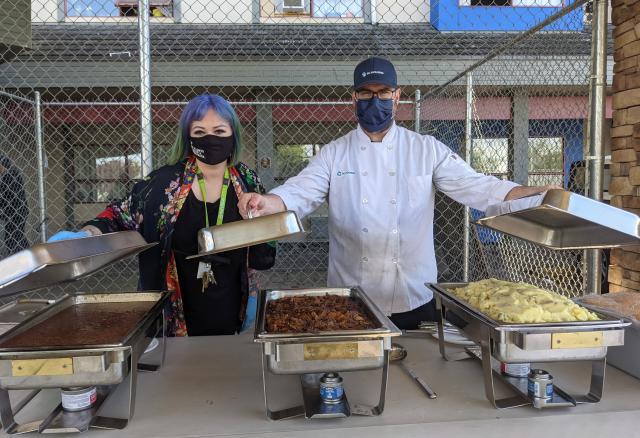 A woman and man wearing masks serve food from behind a table outdoors