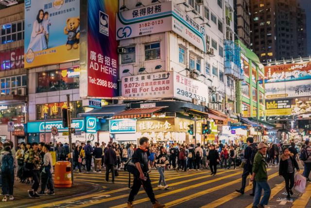 A busy night street scene in Hong Kong, with large groups of people crossing an intersection, backed by well-lit buildings covered with advertisements and signs.