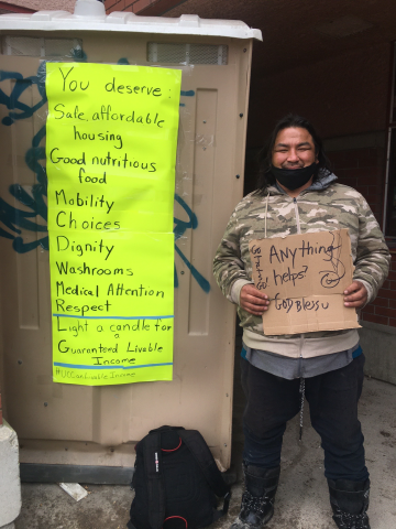 A man with dark skin and rough clothing holding a sign that says "Anything helps," next to another homemade sign listing the benefits of a Guaranteed Livable Income.