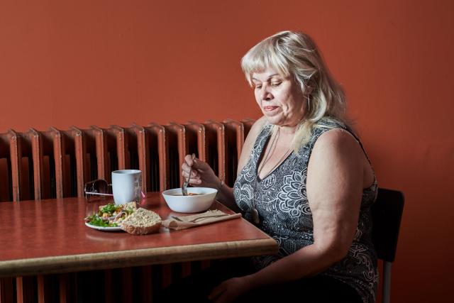 A woman sits at a table eating soup, bread, and a salad against a brown wall.