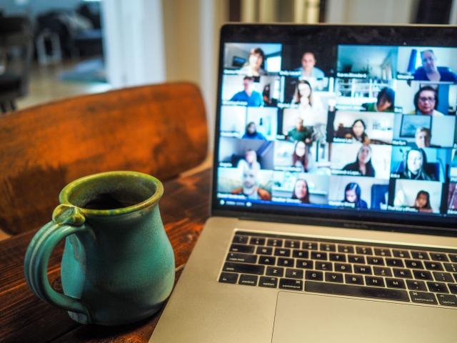 Image: A laptop with tiled images of meeting participants on the screen sits on a desk with a coffee mug beside it