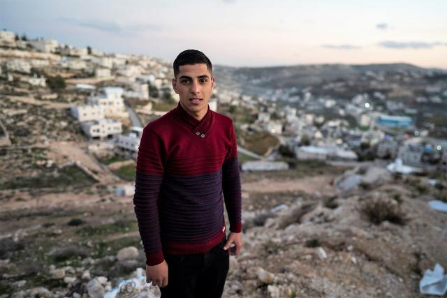 Image: A teenage Palestinian boy wearing a maroon sweater standing in front of a hilly and rocky background.