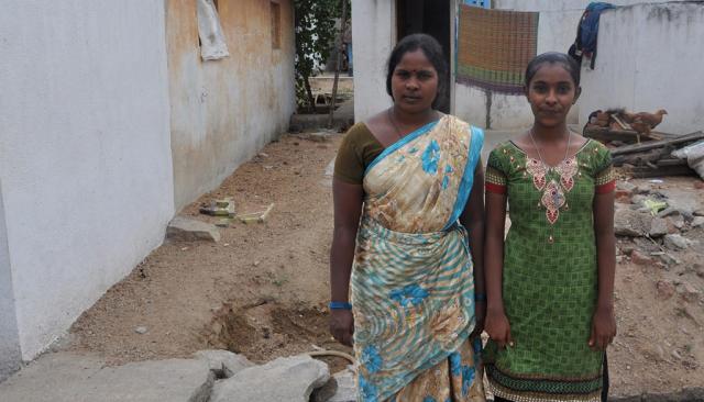 An Indian woman and her teenage daughter stand in front of a simple concrete house.