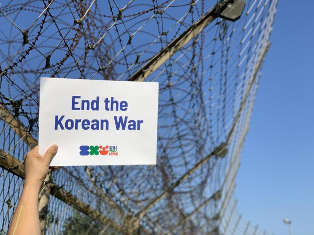 Photo of hand holding up a sign saying End the Korean War with a barbed wire fence in the background