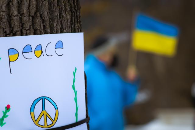 Hand-drawn sign with the word "peace" and the peace symbol in the yellow and blue colours of the Ukrainian flag hangs on a tree.