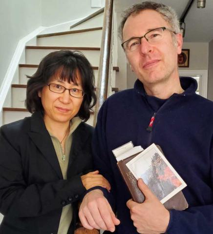 HyeRan Kim-Cragg (left) and husband David Kim-Cragg (right), stand at the foot of a staircase in a house. David is holding a Bible and a postcard of the Diamond Mountains in Korea. Both are wearing black.