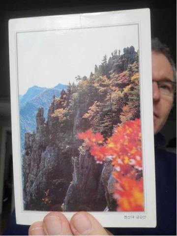 David Kim-Cragg displays an old postcard from Korea showing the Diamond (Kumkang) Mountains, steep, rocky range, dotted with evergreen trees and several deciduous trees which are red and orange as the picture was taken in fall.