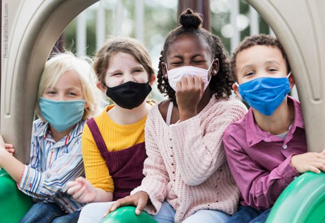 Four young children wearing masks sit together at the top of a playground slide.