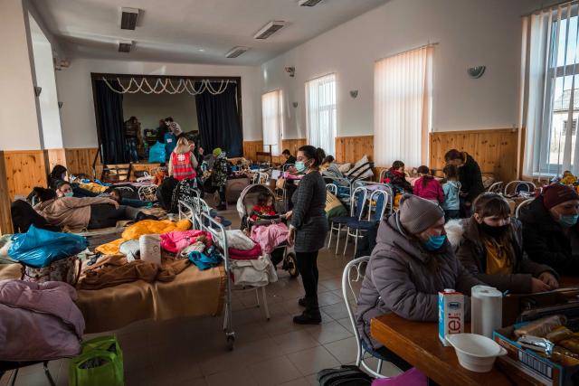 In a crowded refugee centre people sit or stand around beds lined up along the walls.
