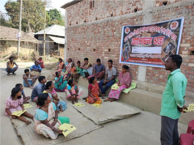 People in India sit on mats on the ground and on a bench along a wall while a man who is standing speaks.