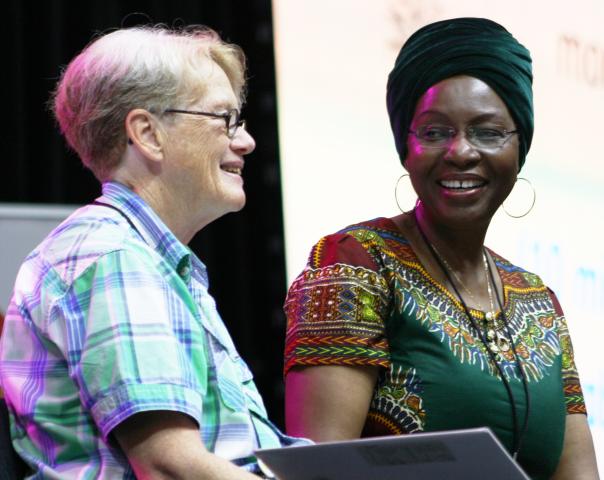 A White and a Black woman sit together talking on a stage at General Council 43.