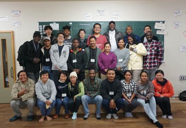 Three rows of young adults from different countries pose in front of a blackboard in a classroom.