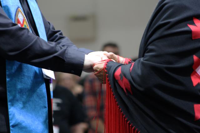 The hands of someone wearing a minister's stole and another person wearing an Indigenous shawl are clasped.