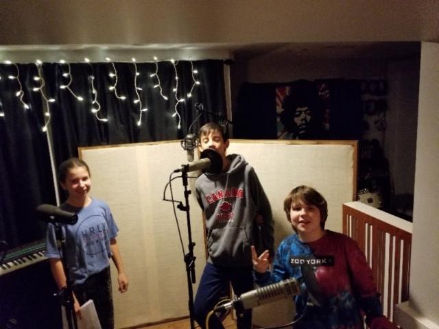 Two boys and a girl, all around 10 years old, stand near a microphone in a recording studio.