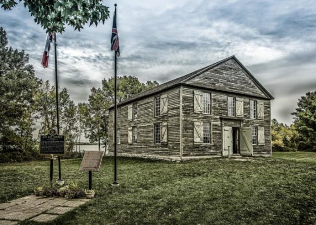 A small church covered in weathered grey clapboard is surrounded by trees, with a historic plaque and the Canadian and Union Jack flags to one side.