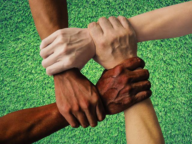 Four hands in different skin colours are clasped at the wrists to form an interlocking square.