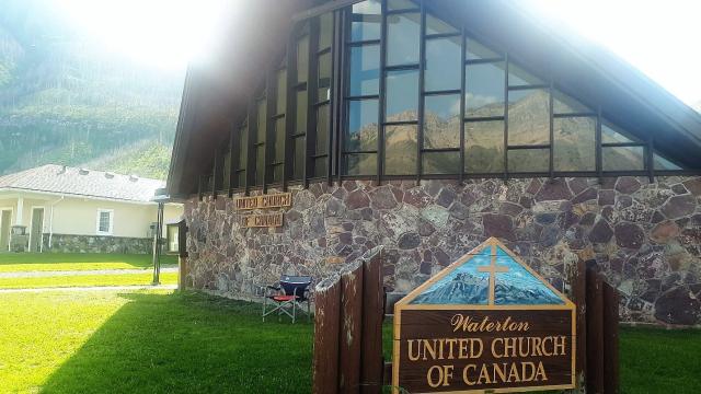 Exterior shot of a church seen against a mountain with a sign in front saying Waterton United Church