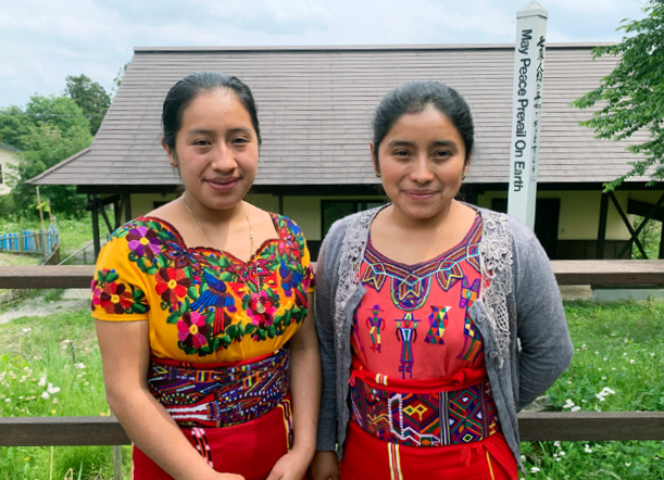 Two young women dressed in Indigenous Guatemalan clothing stand in front of a building in Japan.