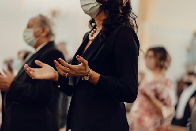 A group of women and men with pandemic masks pray with their palms facing upward at waist level.