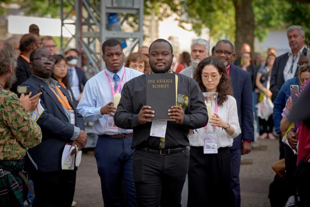 A man carries a large Bible in front of him, followed by a man and woman carrying lit candles, while a crowd watches.