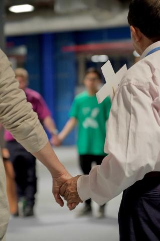 Two hands hold each other, part of a circle of people seen in the background.