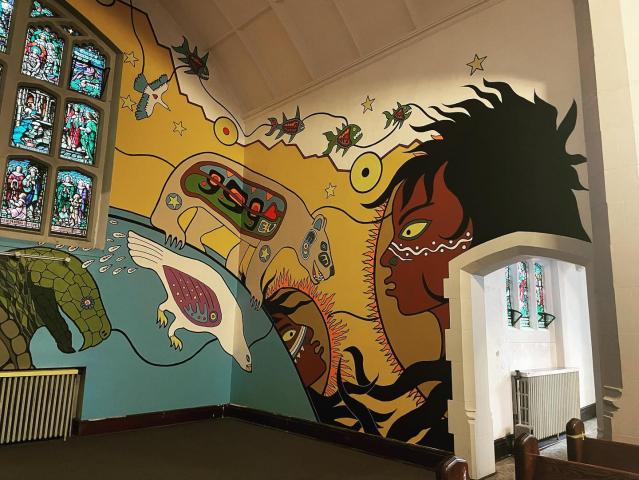 A view of the mural by Indigenous artist Philip Cote showing large First Nations imagery on the interior walls of the church on a yellow background, changing the feel of the sanctuary.