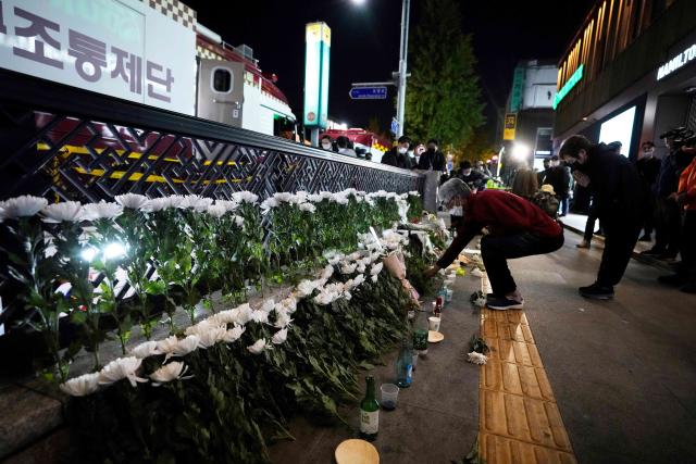 A night scene of an alleyway where a woman places a bouquet with rows of white flowers that form a memorial.