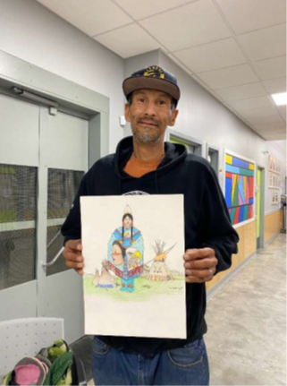 A man wearing a black hoodie and baseball cap stands in a hallway holding his drawing of Indigenous people.