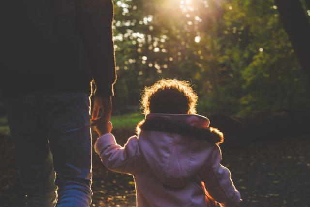 A man holding the hand of his young daughter, who's about 3 years old, walk away from the camera on a forest path. 