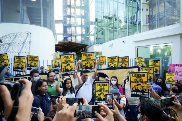 A crowd of people hold up yellow and black signs saying Libertad por los lideres comunitarios in front of an office building.