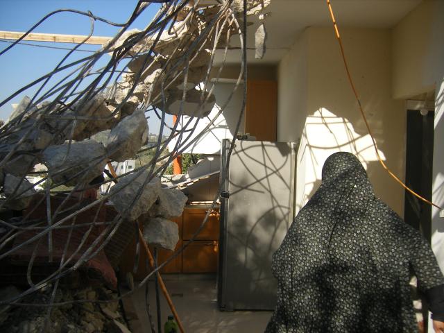 A woman in a chador is seen from behind, walking toward the remains of her home, which had just been demolished. Twisted iron bars and rough concrete pieces hang from what little of the structure remains standing.
