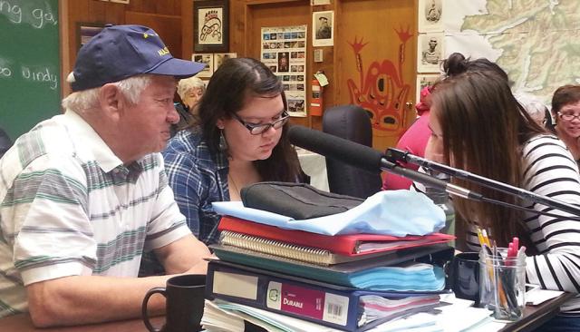 An older man and two young women sit at a desk stacked with papers, books, and binders. In the background are more small groups sitting at tables.
