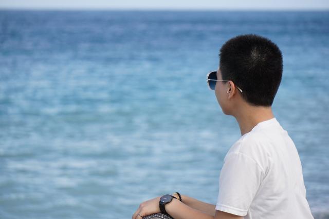 A young person in a white shirt and sunglasses sits on a seawall and looks out to sea on a bright, sunny day against a deep blue sky.