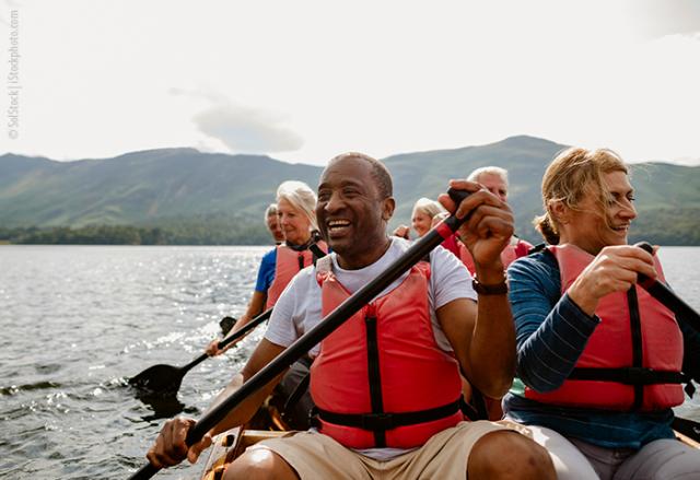 Six adults wearing life jackets paddle a canoe in a lake with mountains in the background.