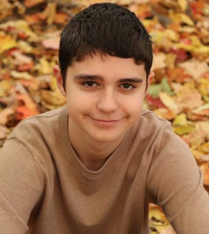 A White teenager with short dark hair sits in fall leaves and looks at the camera with a slight smile.