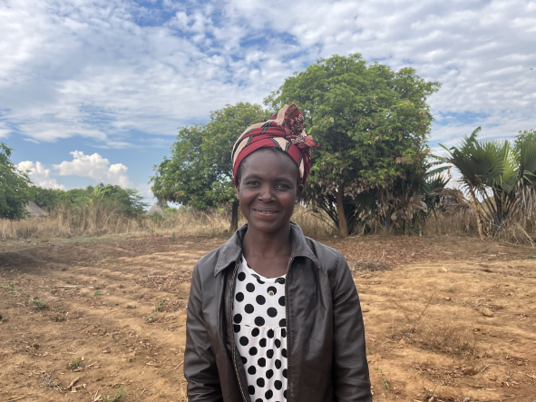 In Zambia, Merit Mabenga, a woman wearing an African headscarf, stands smiling against a background of a field with tilled rows.